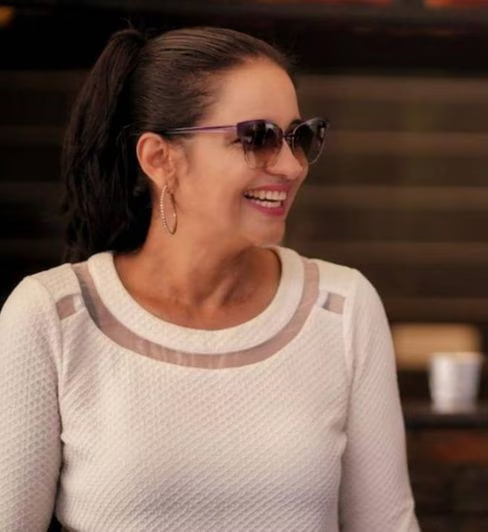 A smiling woman with dark hair and sunglasses, looking to her right.