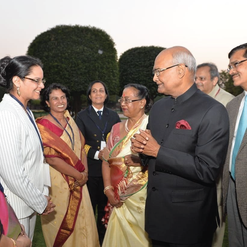 A group of people including a woman in a striped blazer and a man in a formal black suit, at an outdoor event.