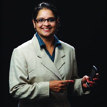 A smiling woman with glasses and a light-colored blazer against a dark background, holding a small object.